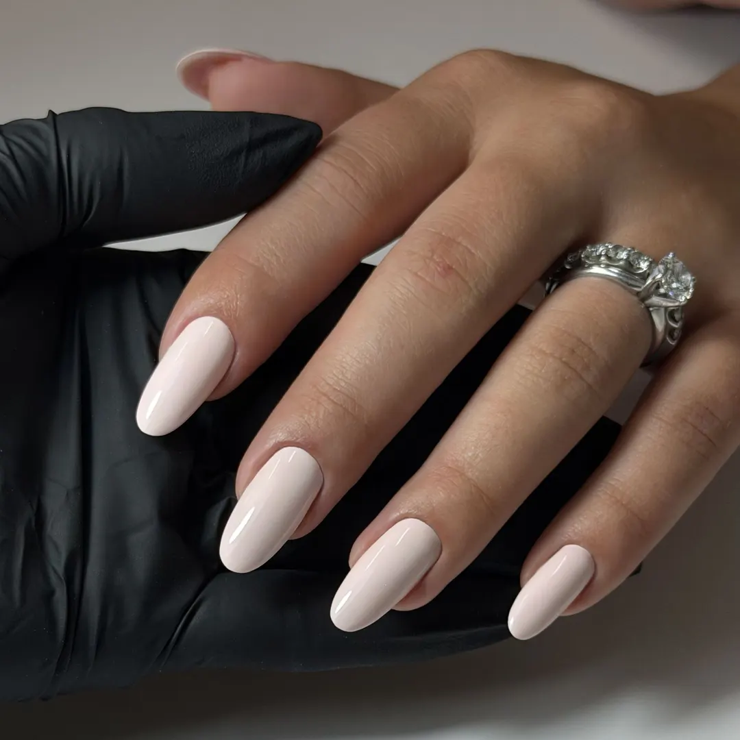 Russian manicure with milky nude gel on almond nails, resting on a technician's black-gloved hand